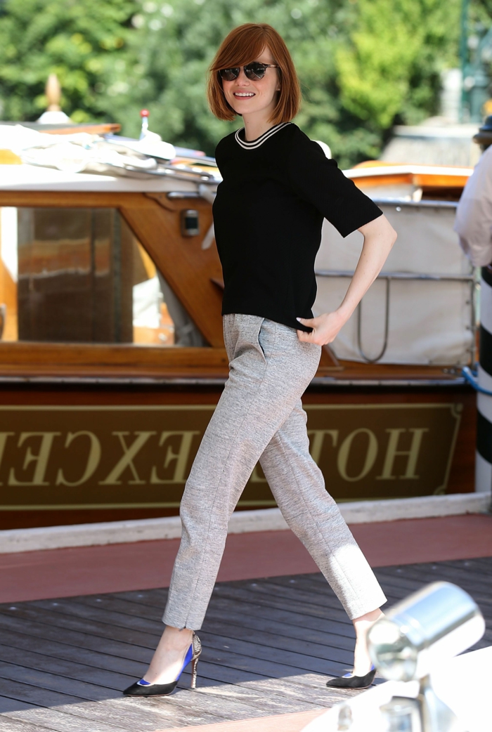 cortes de pelo chica. Emma Stone con peinado melena midi con flequillo hacia un lado,vestida con camisa negra de media manga, pantalon gris y tacones