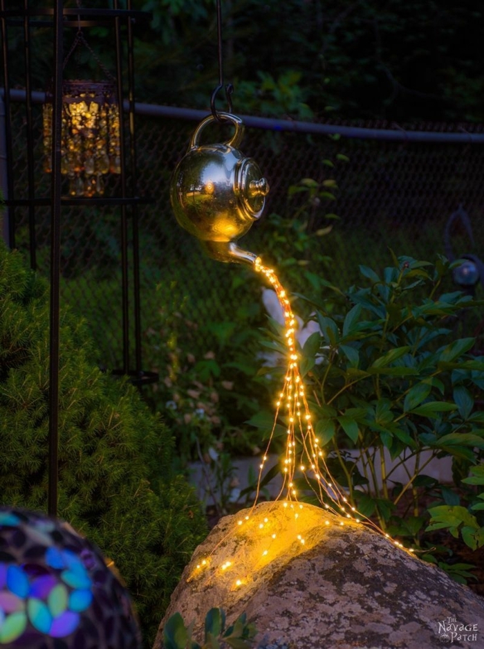 ideas para jardines, caldera colgande de metal con decoración de luces cayendose en una piedra