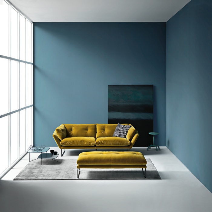 decoracion salon pequeño sofa de doble asiento en color mostaza y taburete alargado, con alfombra en gris y ventanas grandes