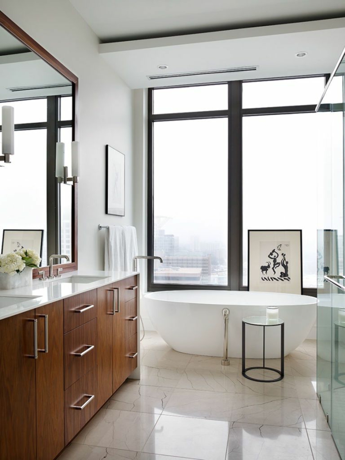 cuarto de baño de diseño decorado en colores claros, suelo con baldosas de mármol, bañera exenta oval y grandes ventanales 