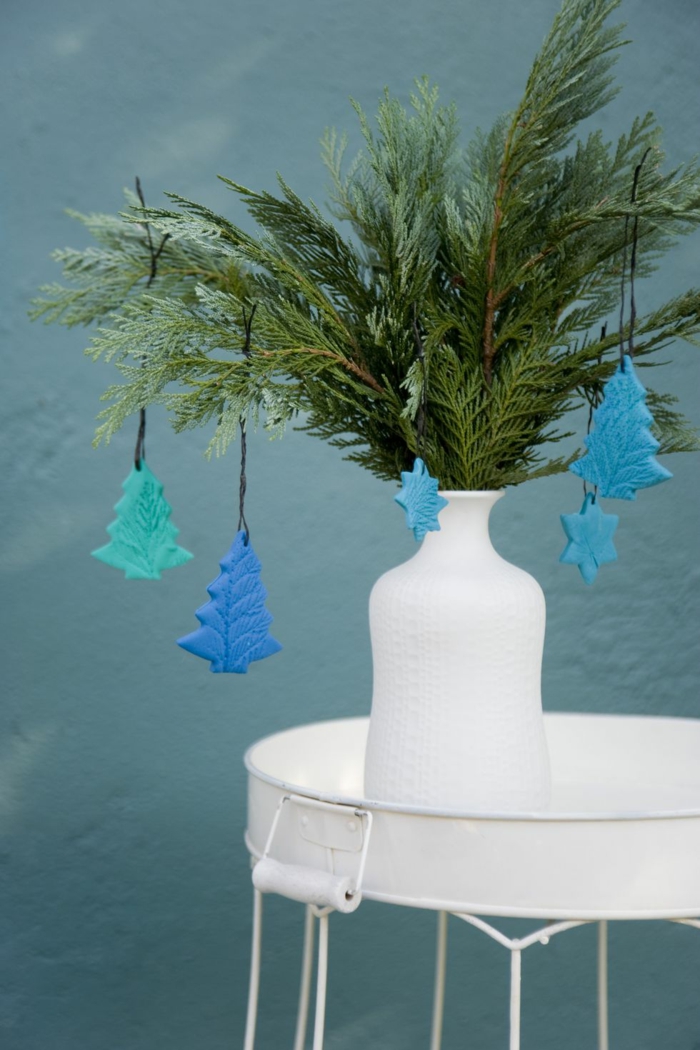 decoracion casera con ramas de pino, florero blanco en una mesa blanca estilo minimalista, pequeños adornos de arcilla en color azul