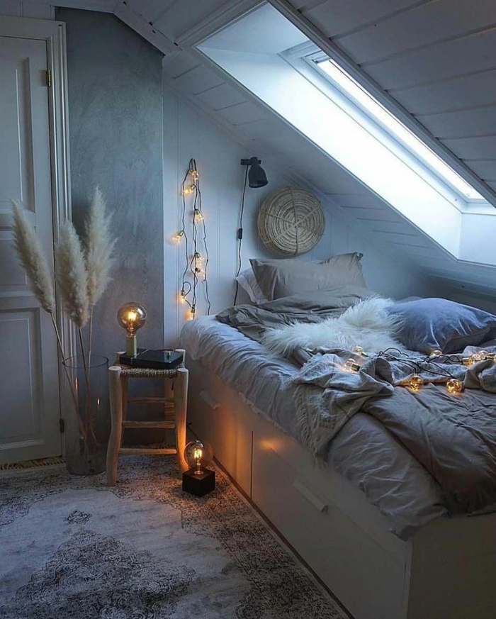 espacio buhardilla con bonito rincón de lectura, cama con sábanas en color gris, alfombra ornamentada y cadena de luces