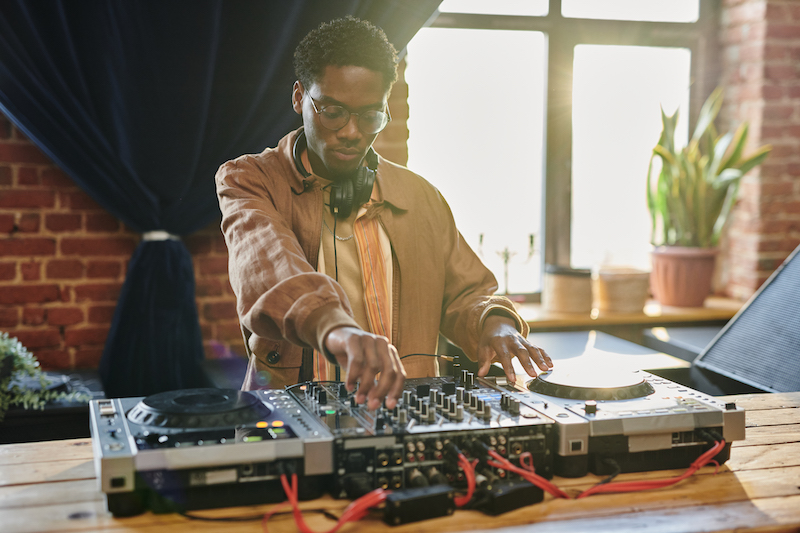 black man in stylish casualwear turning mixers of music equipment